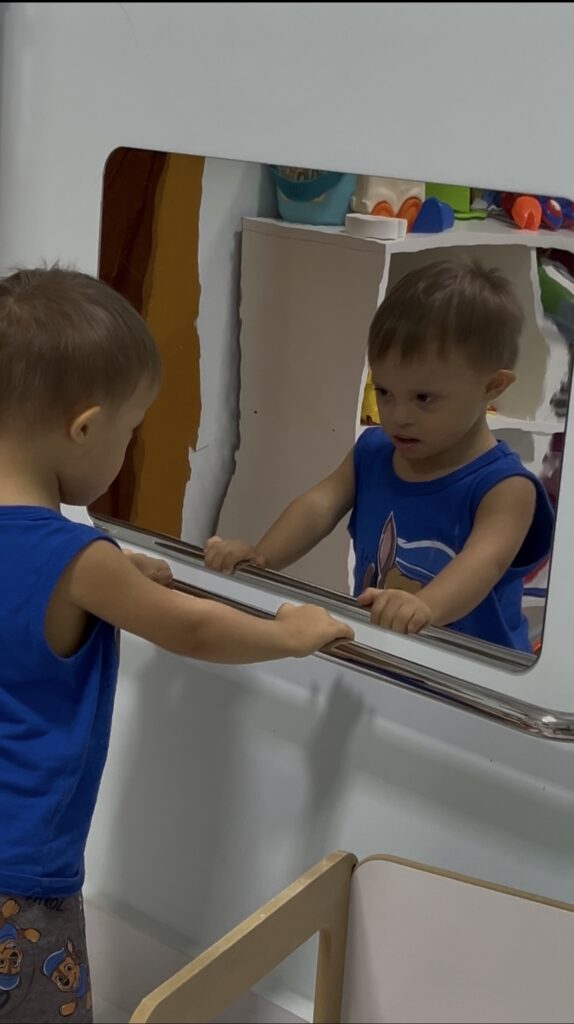 João em pé, segurando a barra de apoio e olhando para o espelho montessoriano na parede